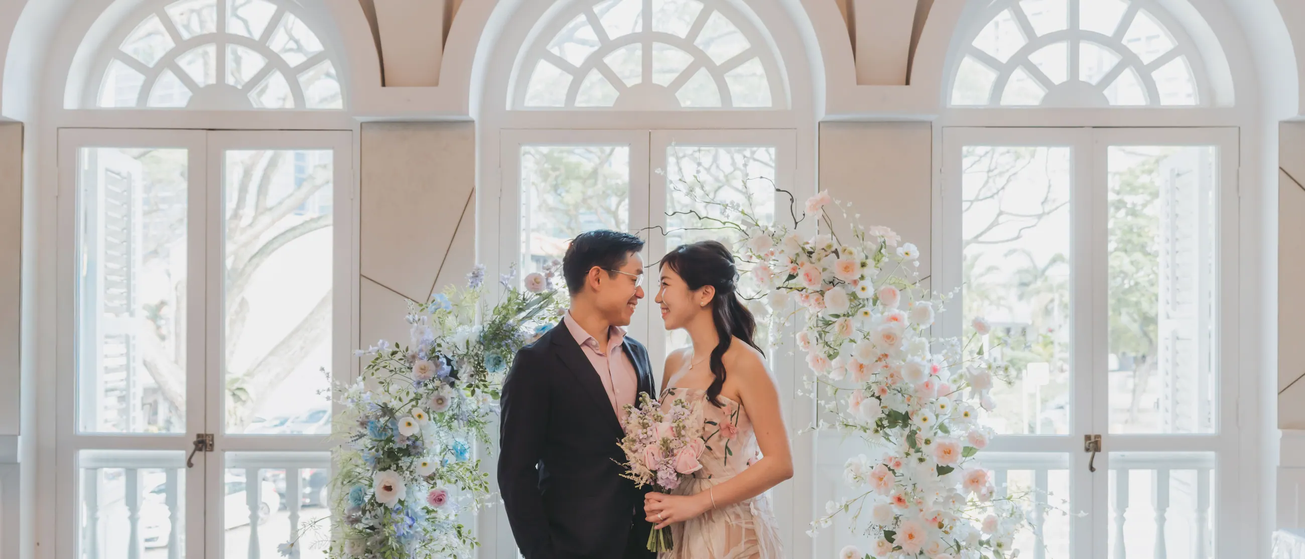 Jingwen and Thai beneath a floral arch in front of tall arched windows