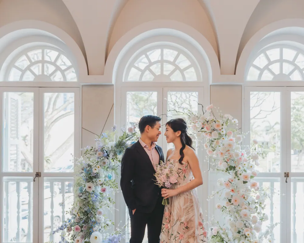 Jingwen and Thai beneath a floral arch in front of tall arched windows