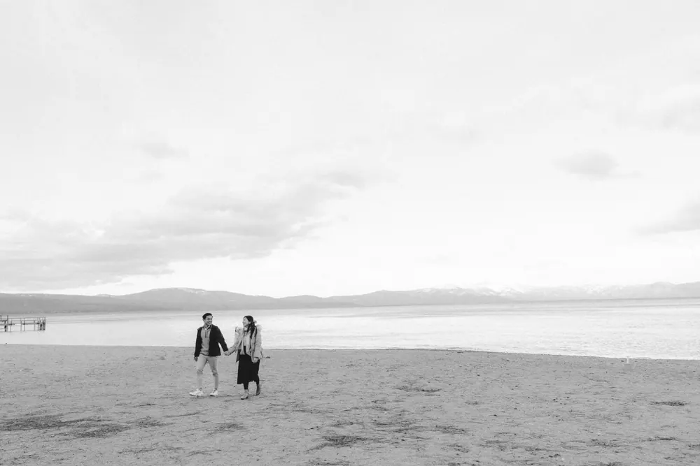 Jingwen and Thai walking hand in hand along the empty shore of Lake Tahoe, mountains across the water